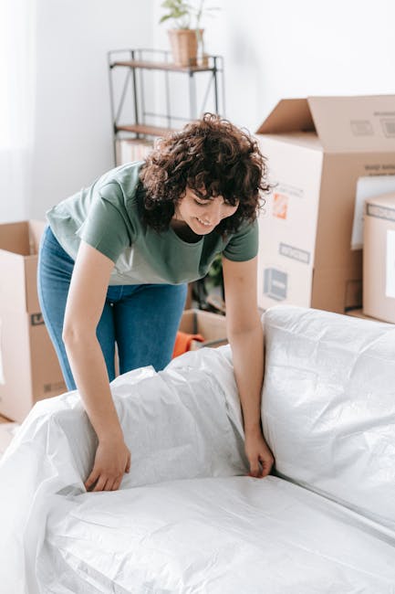 A woman with curly dark hair wearing a green t-shirt and blue jeans is in a well-lit, unfurnished room during a home relocation process. She is carefully wrapping or unwrapping a large piece of furnit