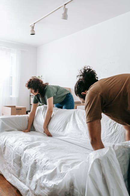A woman with curly dark hair wearing a green t-shirt and blue jeans is in a well-lit, unfurnished room during a home relocation process. She is carefully wrapping or unwrapping a large piece of furnit