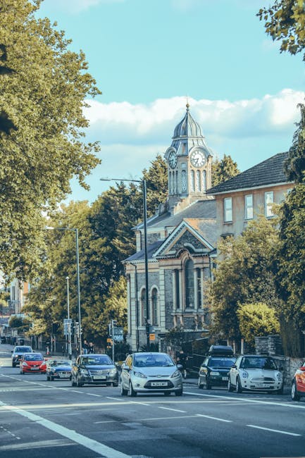 A city street scene featuring a row of parked cars along the pavement with various models and colours situated in front of a historic building with detailed brickwork and arched windows. Behind the building, there is a tall clock tower with a domed roof and a clock face displaying the time, set against a bright blue sky with scattered white clouds. Large leafy trees line the street, providing shade and greenery, and a pedestrian sidewalk runs parallel to the parked vehicles. The overall setting suggests a typical urban area where house removals and furniture transport services could be conducted, with Edgware Movers operating within such a neighbourhood, facilitating home relocation and packing and moving tasks efficiently.
