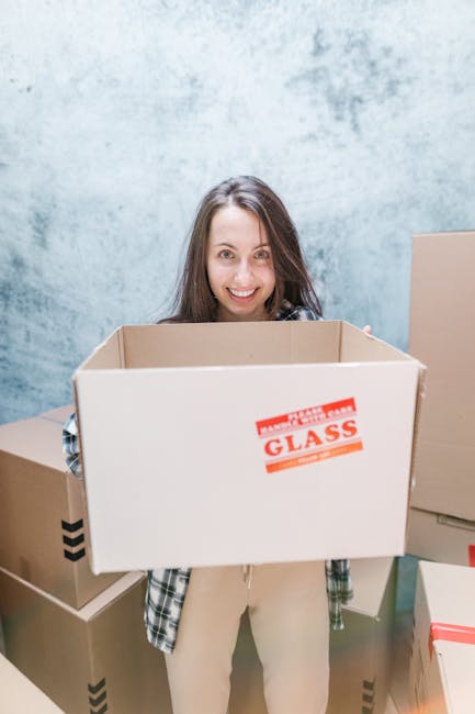 A young woman with long, dark hair smiling while holding an empty cardboard moving box labeled 'FRAGILE GLASS'. She is standing indoors against a textured, light blue wall. Around her are several other cardboard boxes of various sizes, some sealed with packing tape and others open, with a few containing packing materials like bubble wrap or paper. The scene depicts a packing and home relocation process in progress, with the woman preparing for furniture transport or a house move as part of a professional removals service provided by Edgware Movers, indicated by the context of the image and the environment.