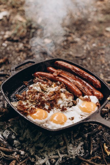 A cast iron skillet placed directly on the ground outdoors, with a rustic dirt and ash background. Inside the skillet, there are three cooked sausages with browned skins, partially covered by melted fat or juices. Next to the sausages, there are two sunny-side-up eggs with firm whites and runny yolks, arranged side by side. Additionally, there is a mixture of crispy bacon pieces or fried minced meat scattered around the eggs and sausages. The scene suggests a home relocation or outdoor cooking activity, reflecting a casual, rustic meal preparation by Edgware Movers’ team during a house removal or moving service, with the cooking equipment and food items serving as visual cues for preparing or enjoying food during a move.