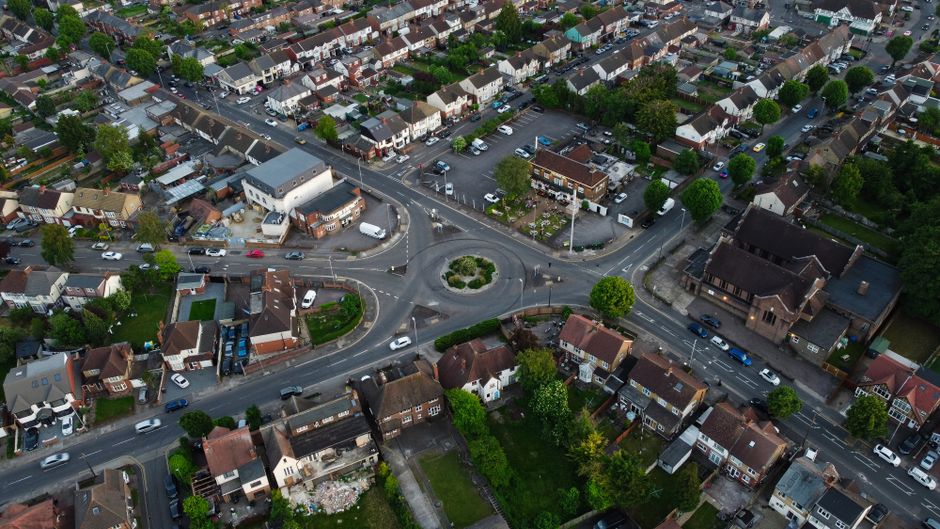 Aerial view of a residential area showing a roundabout surrounded by houses, trees, and parked cars in Burnt Oak and Broadfields Estate, Edgware. The streets are lined with detached and semi-detached homes, some with front gardens and driveways. The roundabout has a small landscaped central island with greenery and shrubs. Several vehicles, including cars and vans, are visible, with some parked along the roads and others approaching or leaving the roundabout. The environment is well-lit with natural daylight, highlighting the orderly arrangement of the neighbourhood and the presence of a home relocation or moving process as part of a house removals service by Edgware Movers. The scene captures the typical layout of a suburban street, suitable for describing furniture transport, packing, and loading activities involved in professional removals.