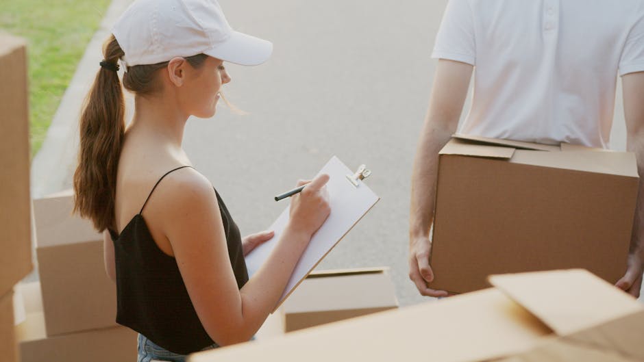 A young woman with light skin, wearing a white cap and a black sleeveless top, is seen holding a clipboard and pen while taking notes during a home relocation process. She stands outdoors near a property, surrounded by several cardboard boxes, some sealed with packing tape and others partially opened, which contain various household items. Her long brown hair is tied back in a ponytail. A person, whose face is not visible, is holding a medium-sized cardboard box filled with household items, positioned next to the woman. The scene appears to be part of a furniture transport and packing operation involving a professional moving service, such as Edgware Movers, with a focus on loading or loading process. The environment is well-lit and suggests an organized activity related to house removals, with items stored on a paved surface or driveway near the property.