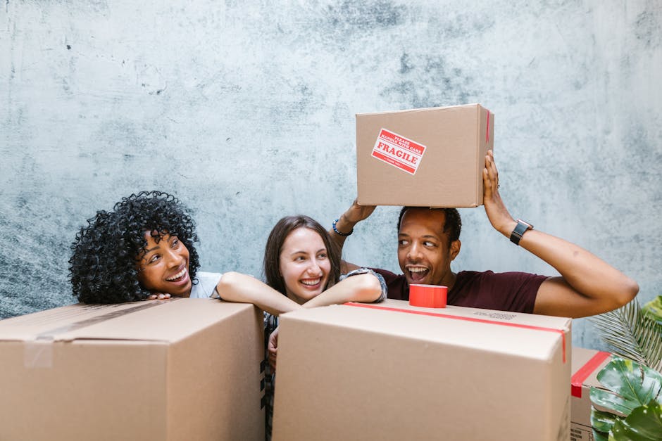 Three individuals in an indoor setting surrounded by cardboard moving boxes, some labelled with fragile stickers, as part of a home relocation process. The person on the right is holding a medium-sized cardboard box labeled 'Fragile' above their head, while the other two are engaging in the loading process, with one person resting their chin on a box and another smiling in the middle. The boxes are made of brown cardboard, some wrapped with packing tape, and are positioned on a flat surface, possibly a table or floor, with additional packing supplies like tape visible. The background features a textured grey wall, and natural light illuminates the scene. The image relates to packing and moving activities typical of a house removal, and natural references to furniture transport and loading are contextually relevant for Edgware Movers' services on their website page about avoiding hidden fees during relocations.